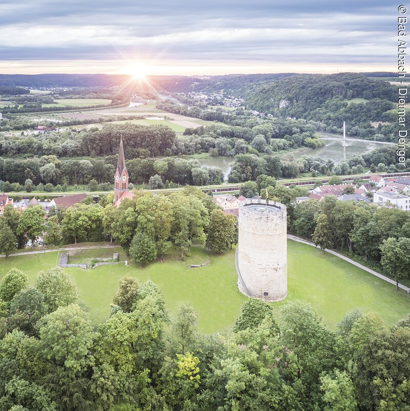 Luftaufnahme einer Landschaft mit einem runden Turm und einer Kirche mit spitzem Turm, umgeben von B&auml;umen und Wiesen.