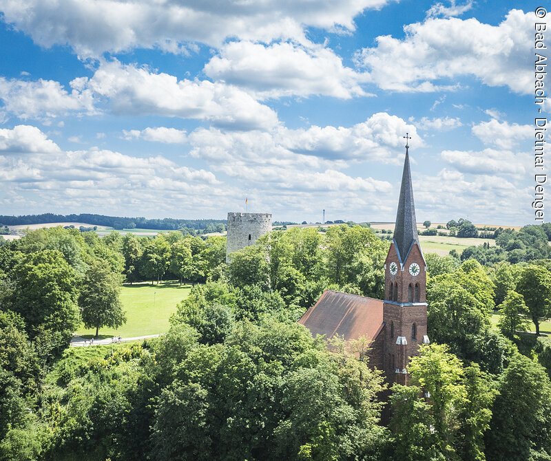 Kirche mit rotem Dach und Turm mit Uhr in gr&uuml;ner Landschaft, daneben ein runder Steinturm.