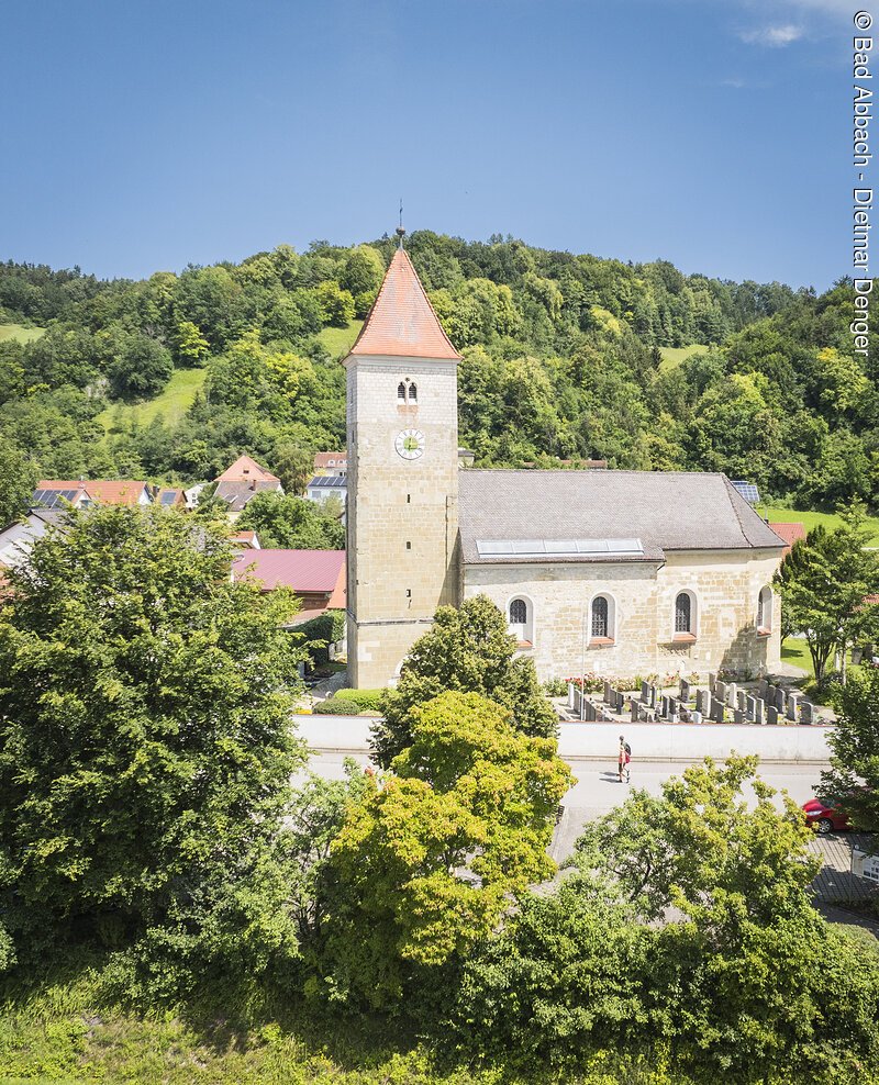 Das Luftbild zeigt die Kirche in Oberndorf aus erh&ouml;hter Perspektive. Die romanisch anmutende Kirche mit quadratischem Turm und rotem Zeltdach steht im Zentrum des Bildes. Umgeben ist sie von einem gepflegten Friedhof mit Grabsteinen und gr&uuml;nem Rasen. Der Himmel ist blau mit leichten Schleierwolken.