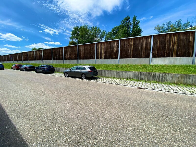 Stra&szlig;e mit parkenden Autos, im Hintergrund L&auml;rmschutzwand und B&auml;ume unter blauem Himmel.