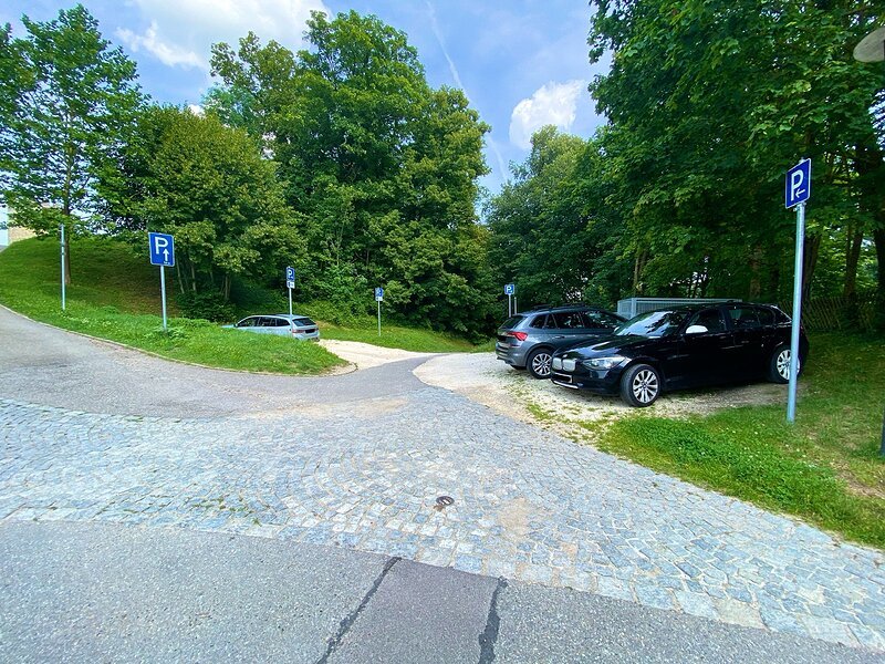 Parkpl&auml;tze auf geschottertem Boden am Waldrand mit mehreren blauen Parkplatzschildern und geparkten Autos.