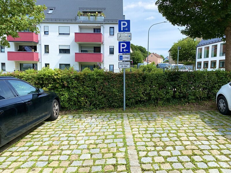 Parkplatz mit Kopfsteinpflaster, zwei Autos am Rand, Hinweisschilder f&uuml;r Parkregelungen vor Hecke und Wohnh&auml;usern im Hintergrund