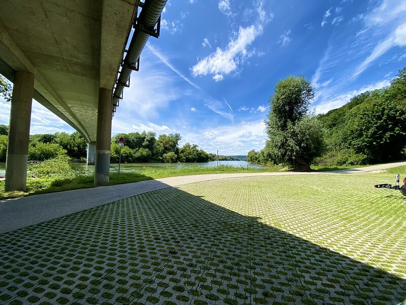 Unter einer Br&uuml;cke mit Pfeilern ein gepflasterter Weg, im Hintergrund Fluss und bewaldete Ufer unter blauem Himmel mit Wolken.