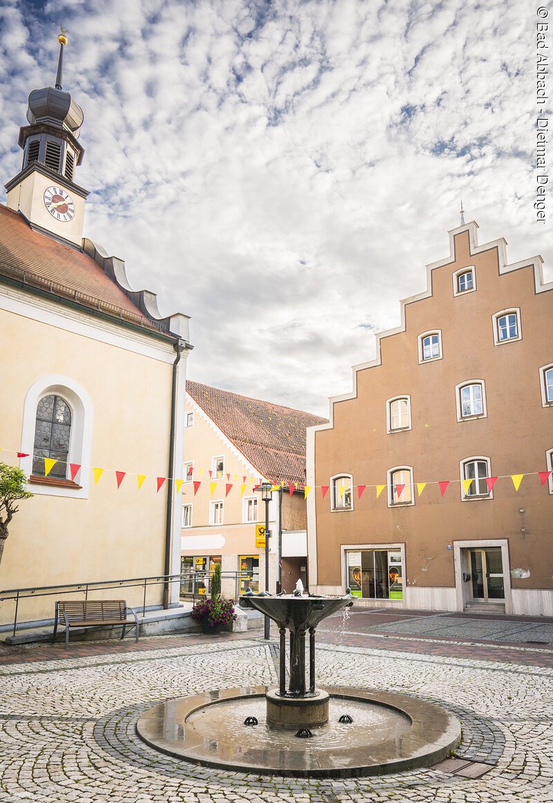 Brunnen zwischen Bürgertreff und Marktkirche Kleiner Platz mit Brunnen, umgeben von historischen Gebäuden, mit bunten Wimpeln geschmückt, unter bewölktem Himmel.
