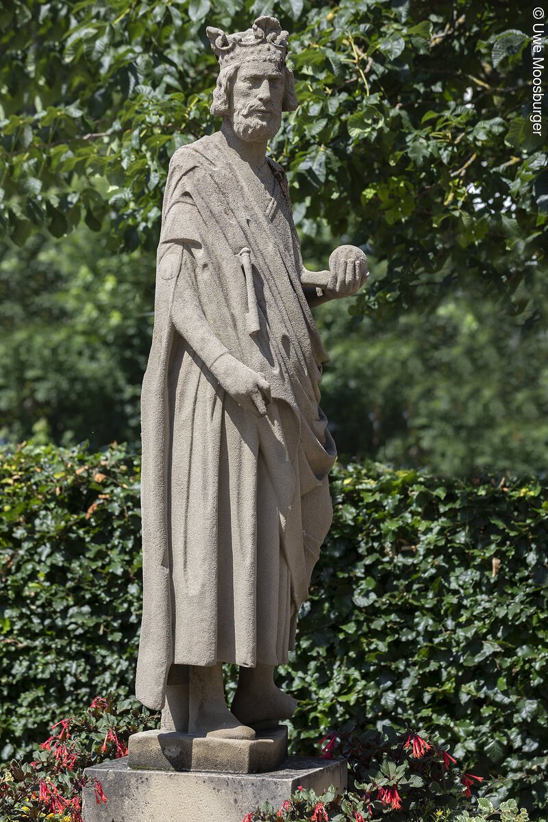 Markt Bad Abbach Figur Kaiser Heinrich II. Steinerne Statue eines bärtigen Königs mit Krone und Kugel in der rechten Hand umgeben von grünen Pflanzen.