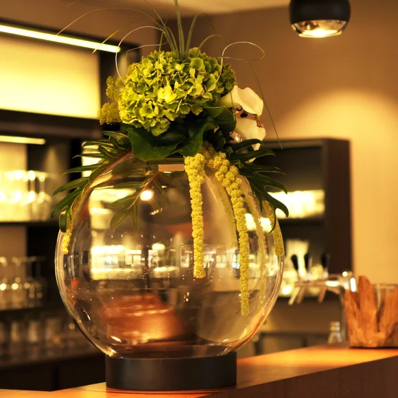 Große Glasvase mit grünen Blumen und Blättern auf einem Tresen in einem warm beleuchteten Raum.