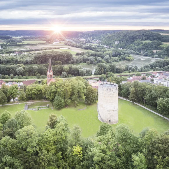 Luftaufnahme einer Landschaft mit einem runden Turm und einer Kirche mit spitzem Turm, umgeben von Bäumen und Wiesen.
