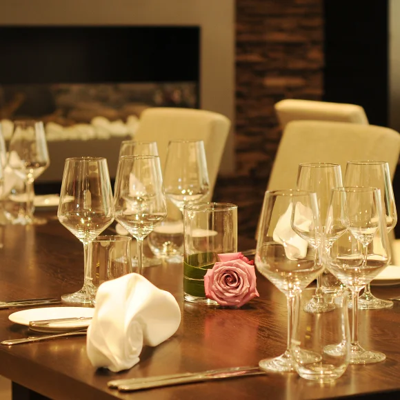 Esstisch mit eingedeckten Gläsern, weißen Servietten und einer rosa Rose in einem Glas auf dunklem Holz.