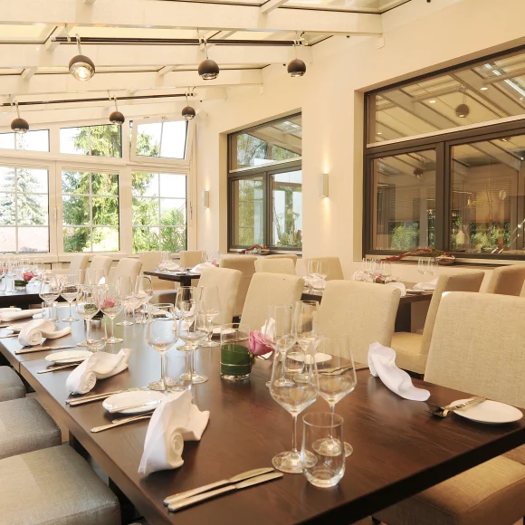 Hell eingerichteter Speiseraum mit gedeckten Tischen und großen Fenstern zu einem Garten hin.