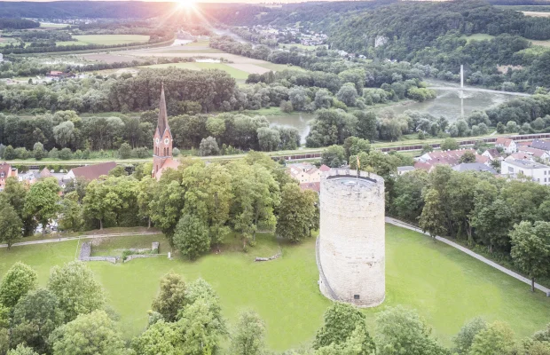 Luftaufnahme einer Landschaft mit einem runden Turm und einer Kirche mit spitzem Turm, umgeben von Bäumen und Wiesen.