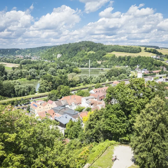 Landschaft mit Fluss, Brücke, Hügeln und einer Stadt mit roten Dächern, umgeben von Bäumen und Wiesen.