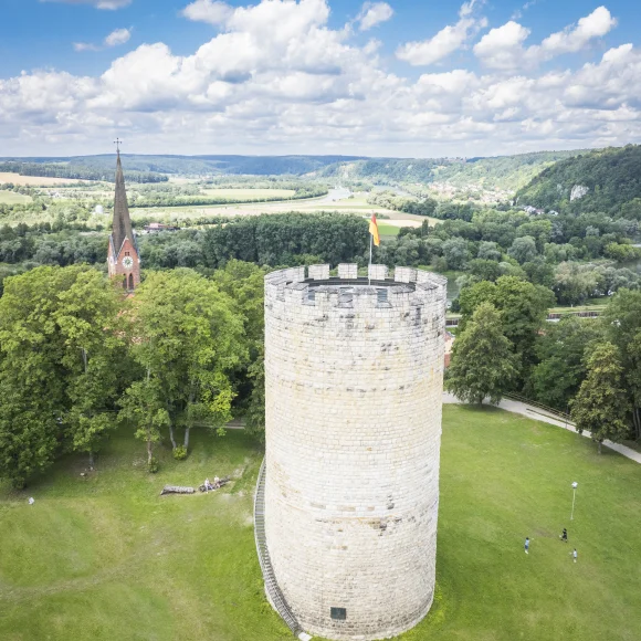 Runder Steinturm mit Fahne auf einer Wiese, im Hintergrund eine Kirche und bewaldete Hügel.