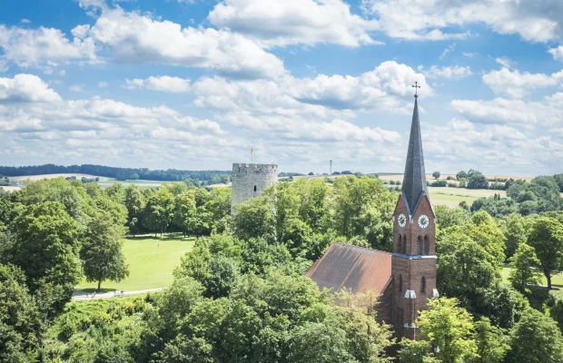 Kirche mit rotem Dach und Turm mit Uhr in grüner Landschaft, daneben ein runder Steinturm.