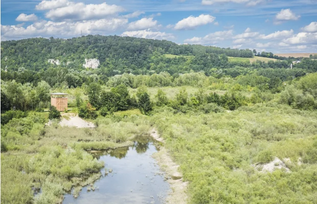 Landschaft mit Fluss, umgeben von Bäumen und Hügeln unter blauem Himmel mit Wolken.