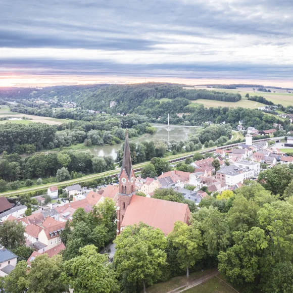 Luftaufnahme einer Stadt mit Kirche, umgeben von Bäumen, Fluss und Hügeln im Hintergrund.