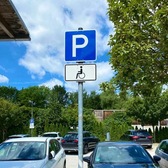 Parkplatzschild mit Rollstuhlsymbol auf einem Parkplatz mit mehreren geparkten Autos und Bäumen im Hintergrund.