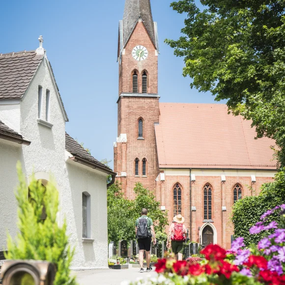 Backsteinkirche mit Turmuhr und spitzem Turm, umgeben von Bäumen und Blumen, zwei Personen gehen auf die Kirche zu.