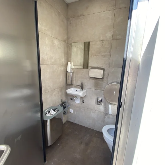 Kleines WC mit grauen Fliesen, kleinem Waschbecken, Spiegel, Papierhandtuchspender, Mülleimer und Toilettenpapierhalter.