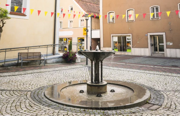 Kleiner Platz mit Brunnen, umgeben von historischen Gebäuden, mit bunten Wimpeln geschmückt, unter bewölktem Himmel.