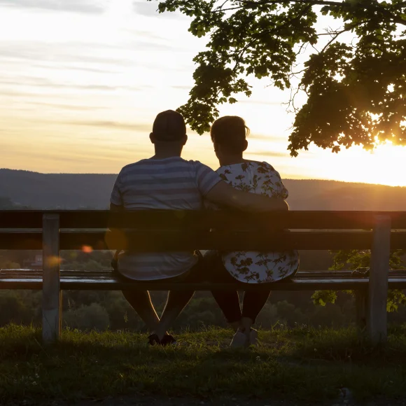 Zwei Personen sitzen auf einer Bank und schauen bei Sonnenuntergang in eine bewaldete Landschaft.