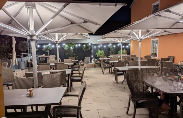 Außenbereich eines Restaurants mit Tischen, Stühlen, großen Sonnenschirmen und Beleuchtung bei Nacht.