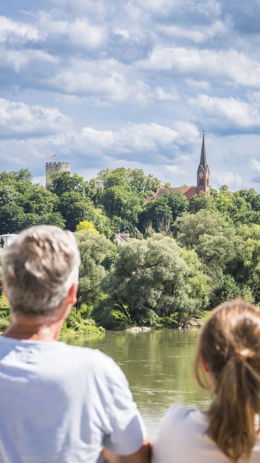 Zwei Personen blicken auf einen Fluss, im Hintergrund eine Kirche und ein Turm auf einem bewaldeten Hügel.