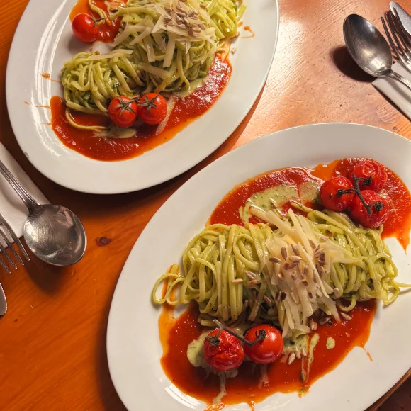 Zwei Teller mit grüner Pasta, Tomatensoße, Kirschtomaten und geriebenem Käse auf Holztisch.