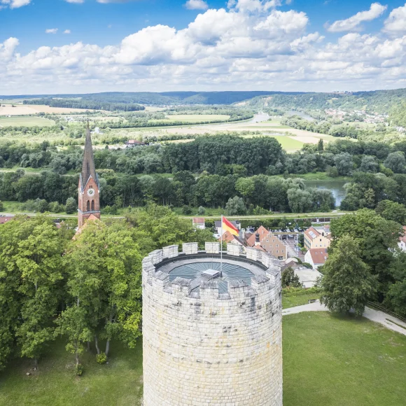 Steinturm mit Flagge, Kirche im Hintergrund, umgeben von Bäumen und Landschaft.