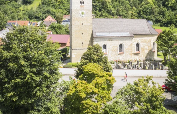 Das Luftbild zeigt die Kirche in Oberndorf aus erhöhter Perspektive. Die romanisch anmutende Kirche mit quadratischem Turm und rotem Zeltdach steht im Zentrum des Bildes. Umgeben ist sie von einem gepflegten Friedhof mit Grabsteinen und grünem Rasen. Der Himmel ist blau mit leichten Schleierwolken.