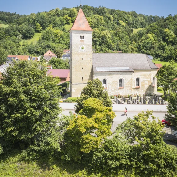 Das Luftbild zeigt die Kirche in Oberndorf aus erhöhter Perspektive. Die romanisch anmutende Kirche mit quadratischem Turm und rotem Zeltdach steht im Zentrum des Bildes. Umgeben ist sie von einem gepflegten Friedhof mit Grabsteinen und grünem Rasen. Der Himmel ist blau mit leichten Schleierwolken.