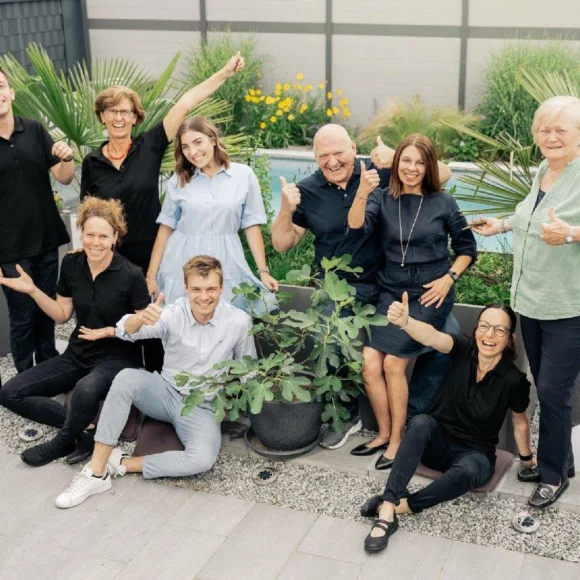 Gruppe von neun Personen steht und sitzt in einem Garten, alle zeigen Daumen hoch, umgeben von Pflanzen.