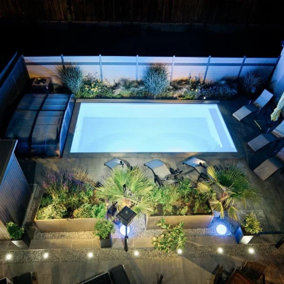 Beleuchteter rechteckiger Pool in einem Garten bei Nacht, umgeben von Pflanzen und Liegestühlen.