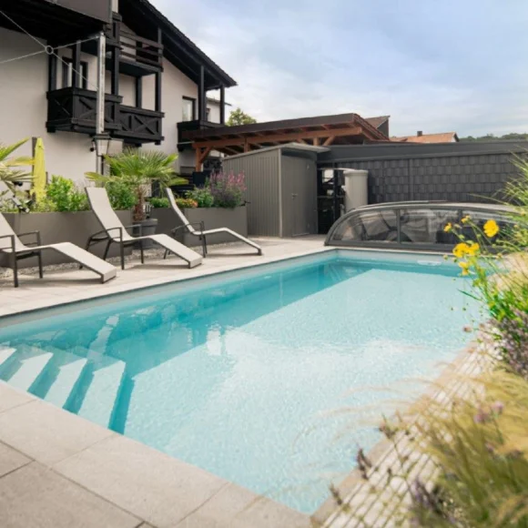 Außenbereich mit rechteckigem Pool, umgeben von Liegestühlen, Pflanzen und einem Gebäude im Hintergrund.