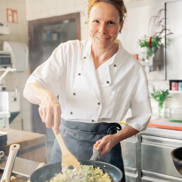 Köchin in weißer Uniform rührt mit einem Holzlöffel in einer Pfanne in einer professionellen Küche.