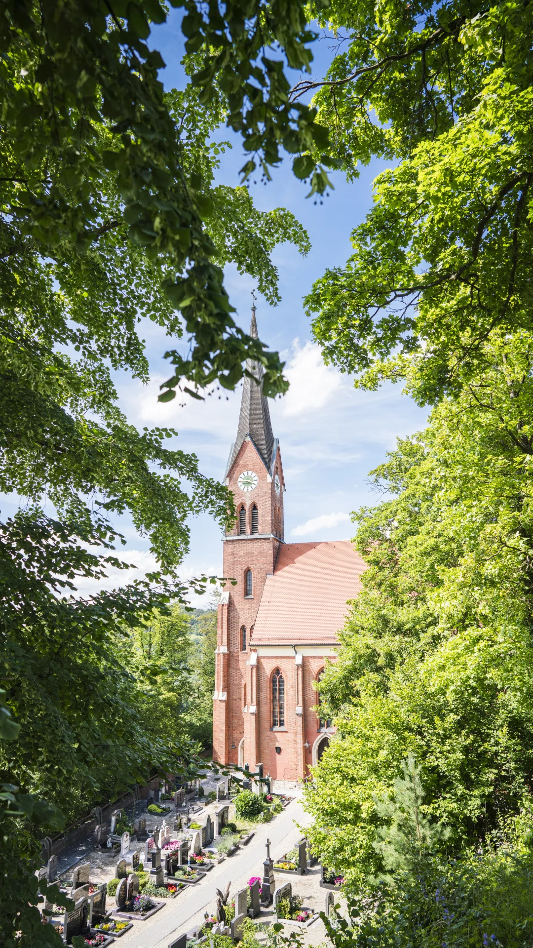 Rote Backsteinkirche mit Turm und Uhr, umgeben von Bäumen und einem Friedhof im Vordergrund.