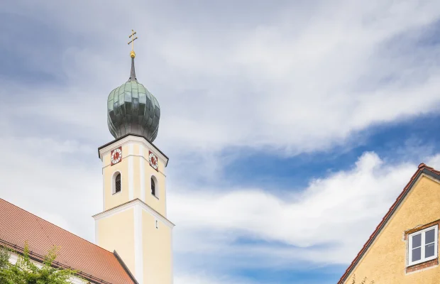 Kirchturm mit grünem Zwiebeldach neben gelbem Gebäude, umgeben von Garten und Zaun, unter blauem Himmel.