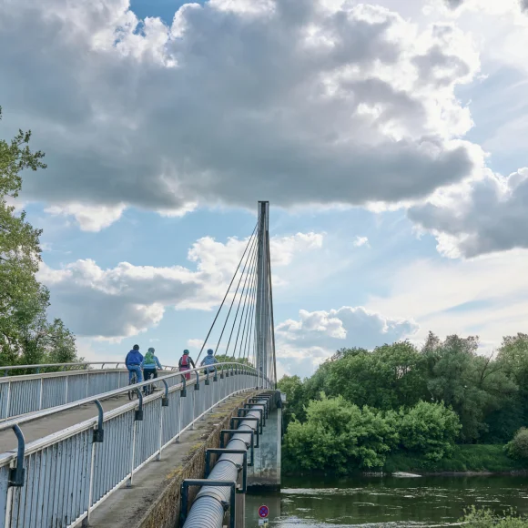 Eine Brücke mit Geländern, auf der Menschen mit Fahrrädern fahren. Darunter fließt ein Fluss, umgeben von Bäumen.