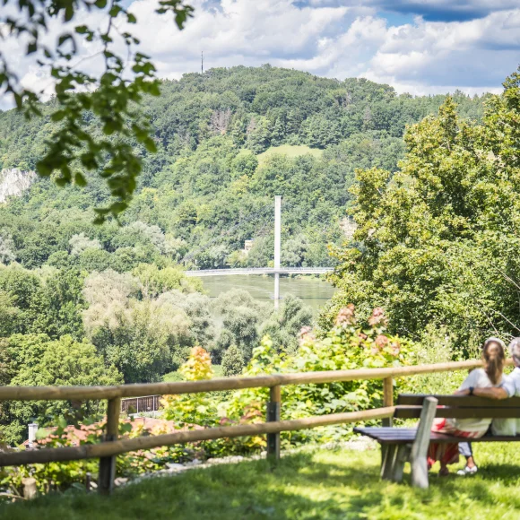 Ein Paar sitzt auf einer Bank mit Blick auf eine bewaldete Landschaft und eine Brücke im Hintergrund.