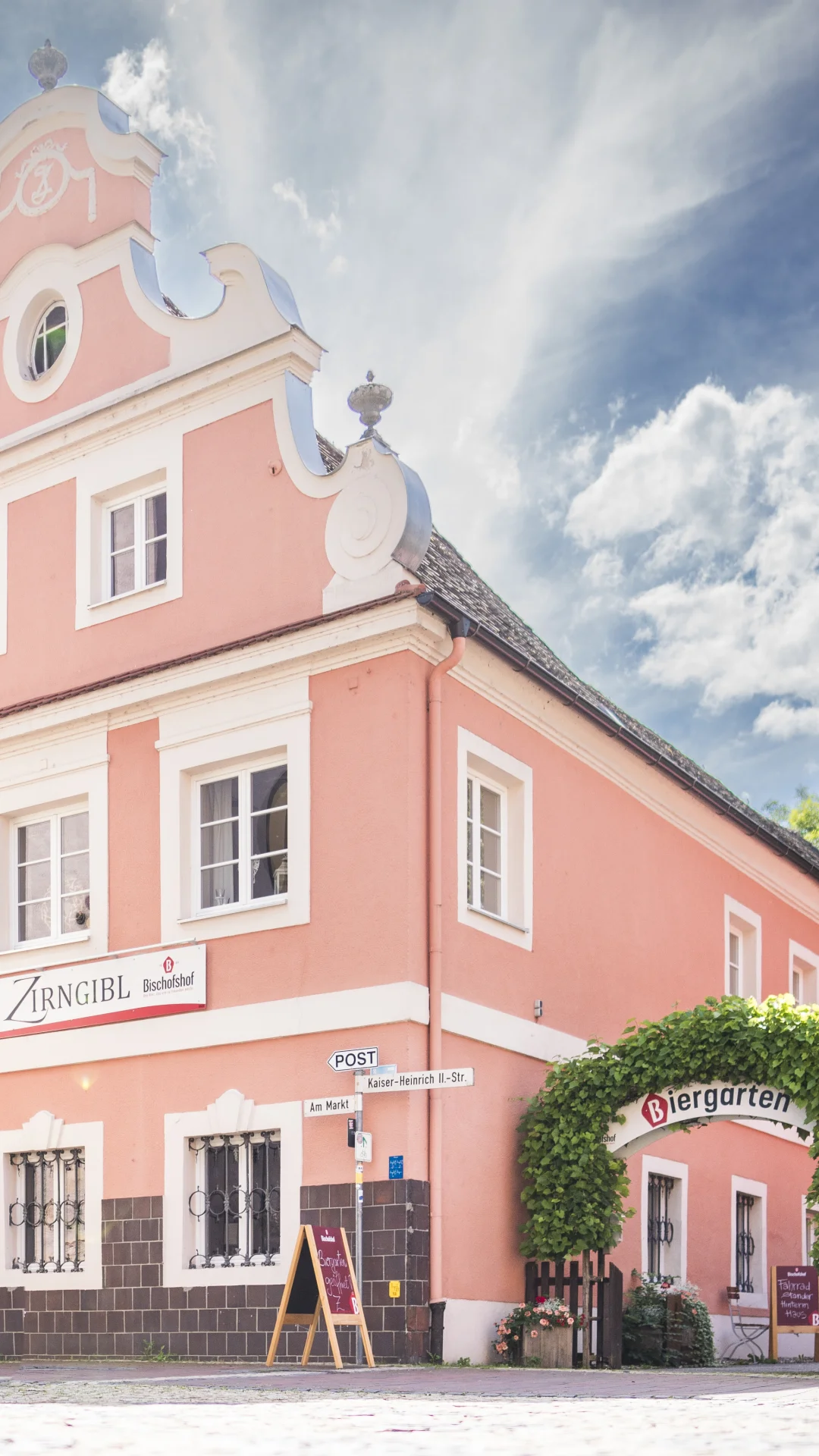 Ein rosa Gebäude mit Barockfassade, daneben ein Biergarten mit grüner Laube, im Vordergrund eine Steinfigur.