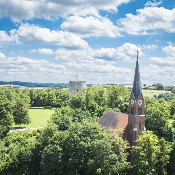 Kirche mit rotem Dach und Turm mit Uhr in grüner Landschaft, daneben ein runder Steinturm.
