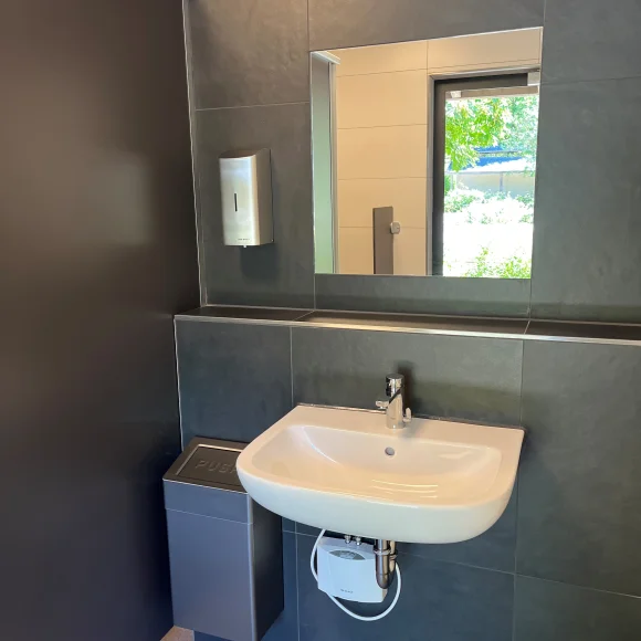 Waschbecken mit Spiegel, Seifenspender und Mülleimer in einem modernen, dunklen Badezimmer mit Fensterblick.