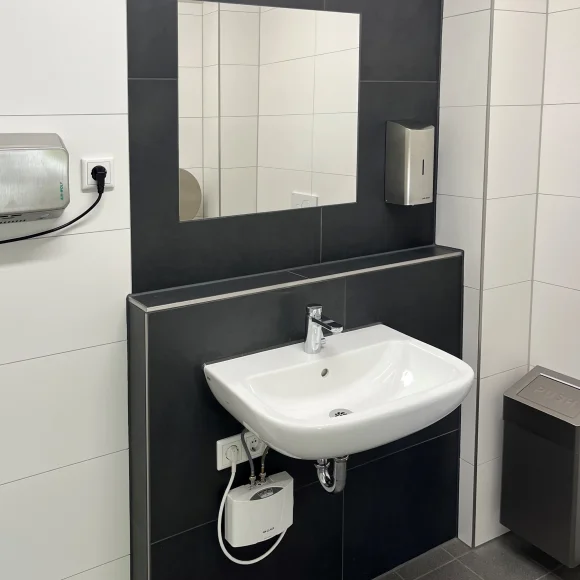 Waschbecken mit Spiegel, Seifenspender und Handtrockner in einem modernen, schwarz-weiß gefliesten Badezimmer.