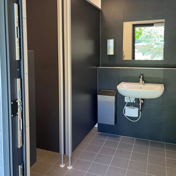 Barrierefreies WC mit grauen Trennwänden, Waschbecken, Spiegel, Seifenspender und Mülleimer.
