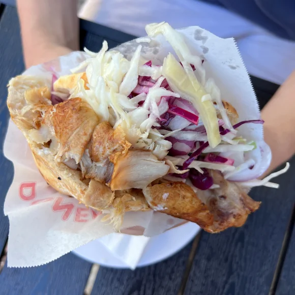 Hand hält gefülltes Fladenbrot mit gegrilltem Hähnchen und Krautsalat in Papierverpackung.